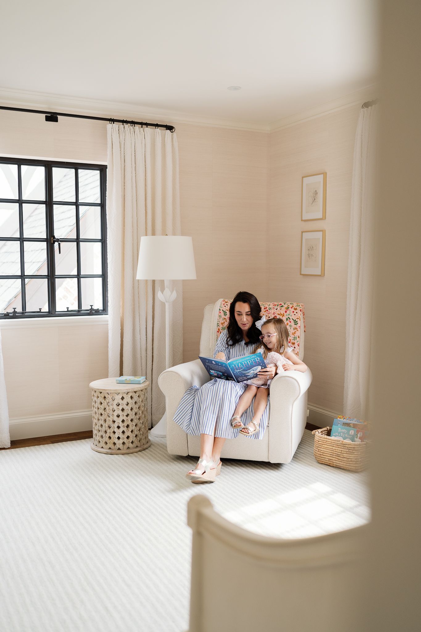 Mother reading a book with young daughter in cozy white nursery chair beside a bright window.