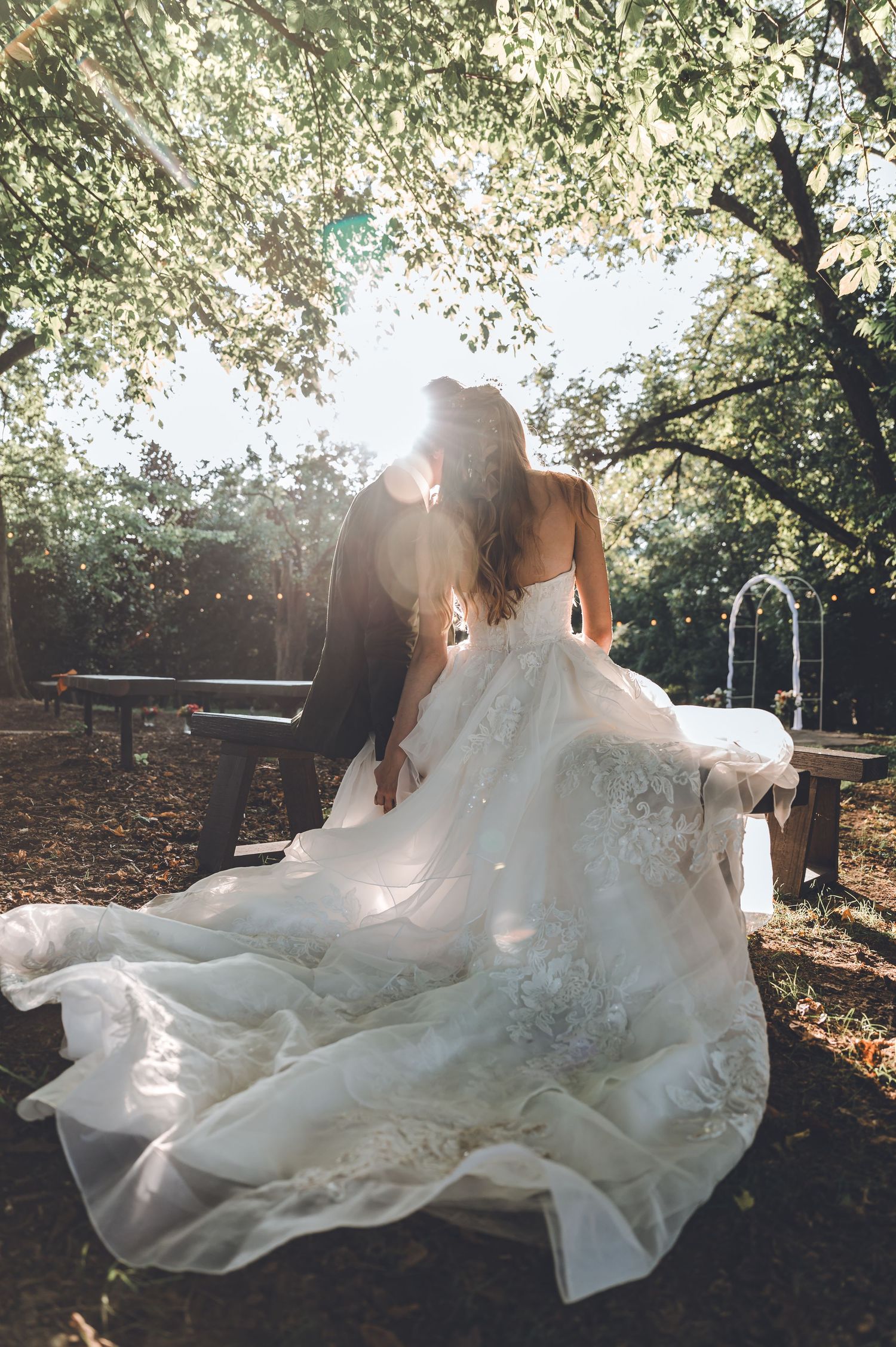 A bride in a flowing lace gown shares a romantic kiss with her groom outdoors under sunlit trees.