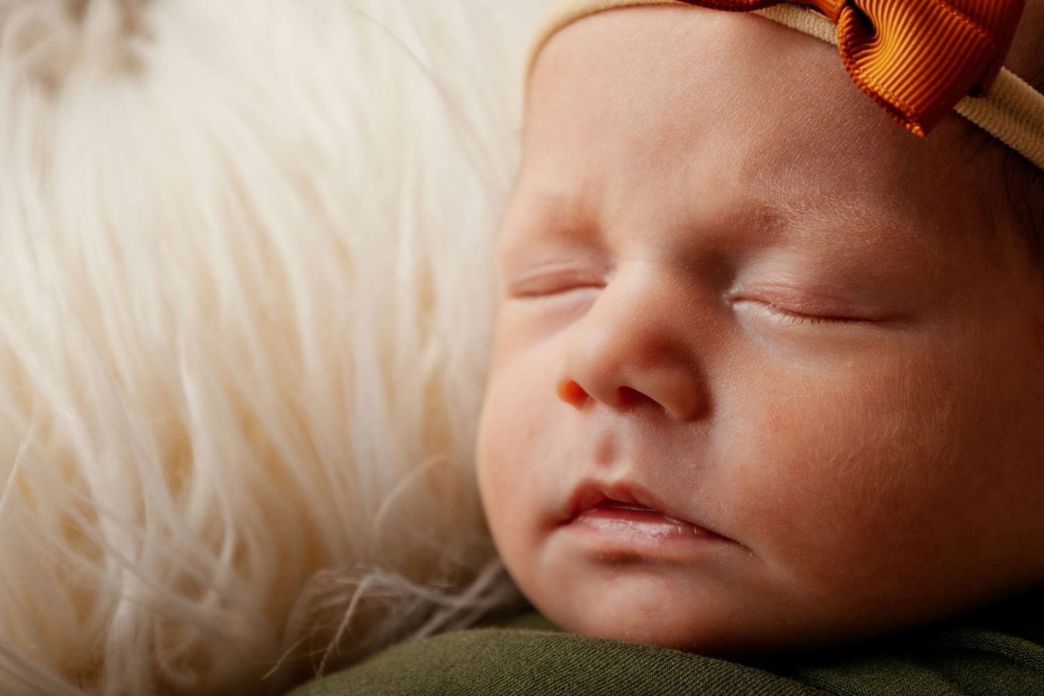 Peaceful newborn baby sleeping with orange bow headband on soft white fur backdrop.