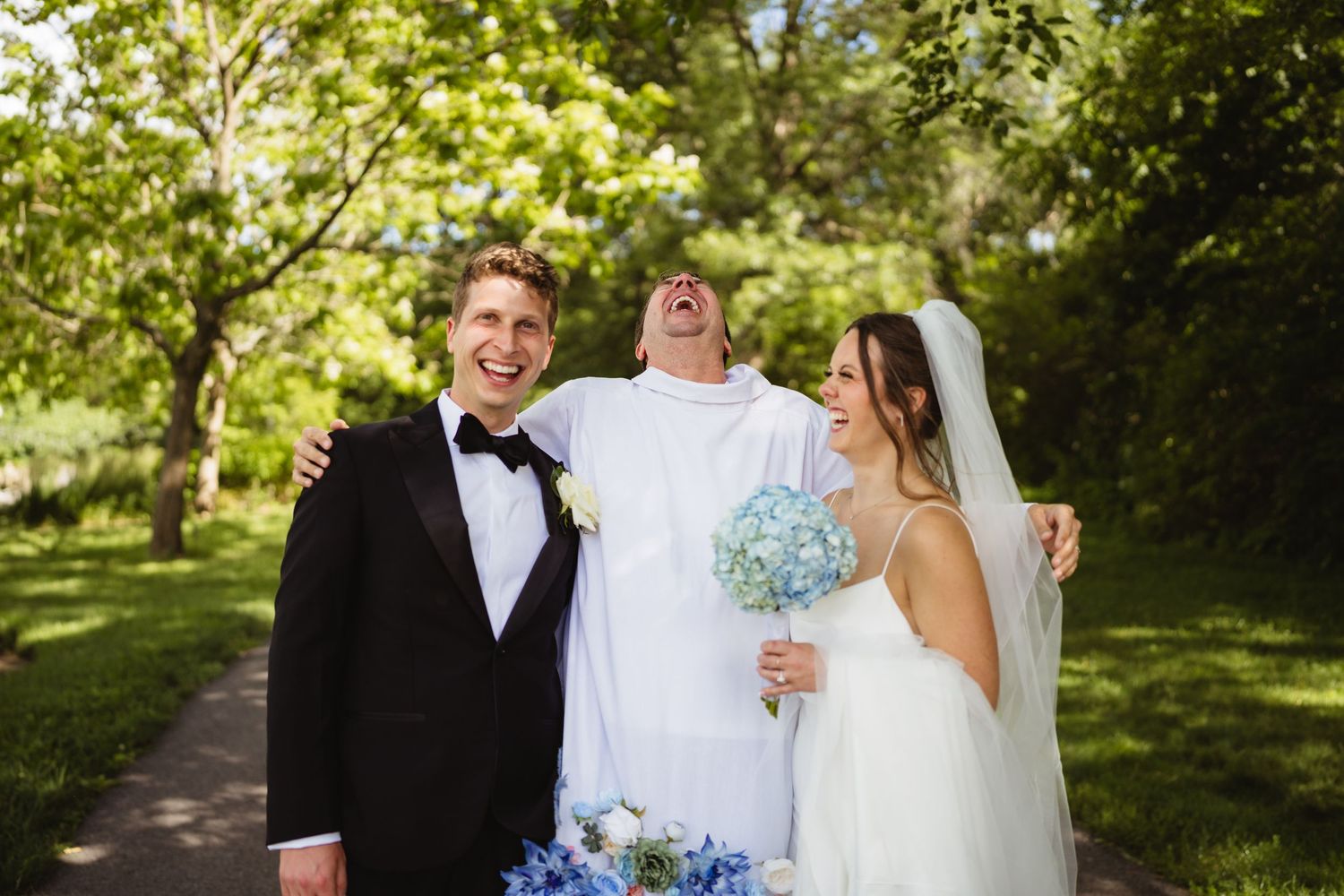 Joyful wedding party with groom in tuxedo, laughing officiant in white robes, and bride holding blue hydrangeas outdoors.