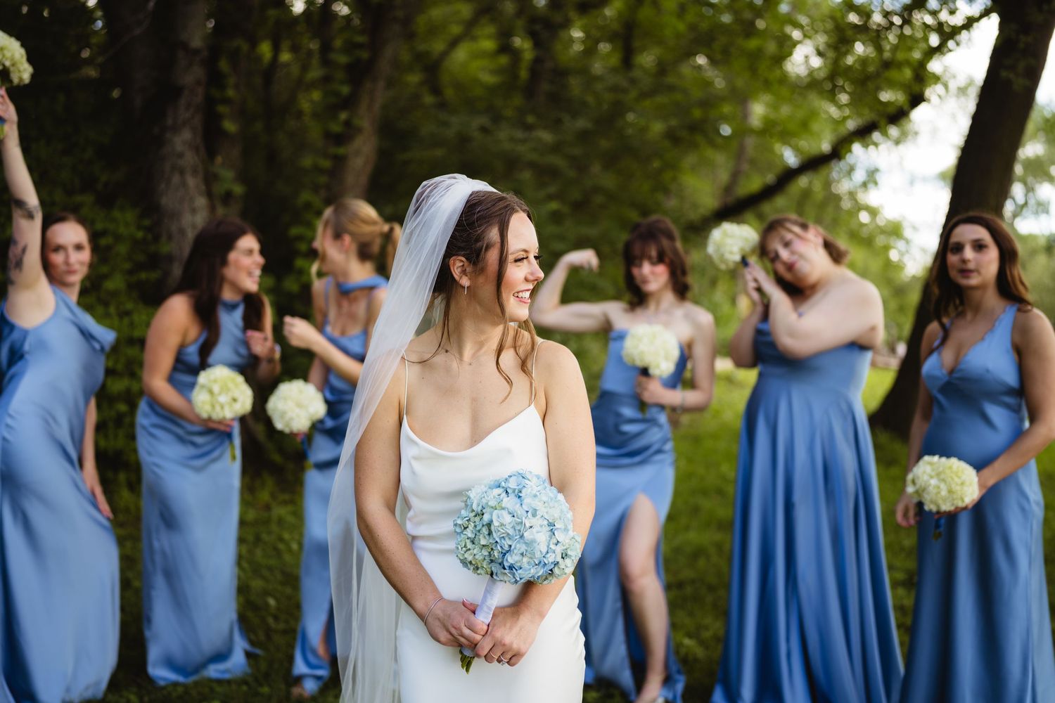 Bride in white gown holds blue bouquet while bridesmaids in blue dresses pose playfully outdoors.
