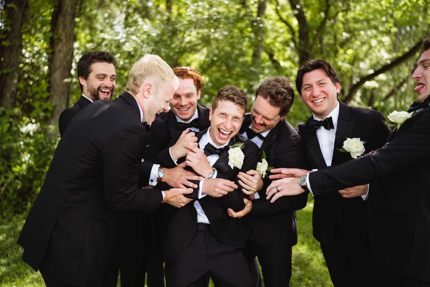 Groom surrounded by groomsmen in black tuxedos laughing outdoors with lush green trees in background at wedding.