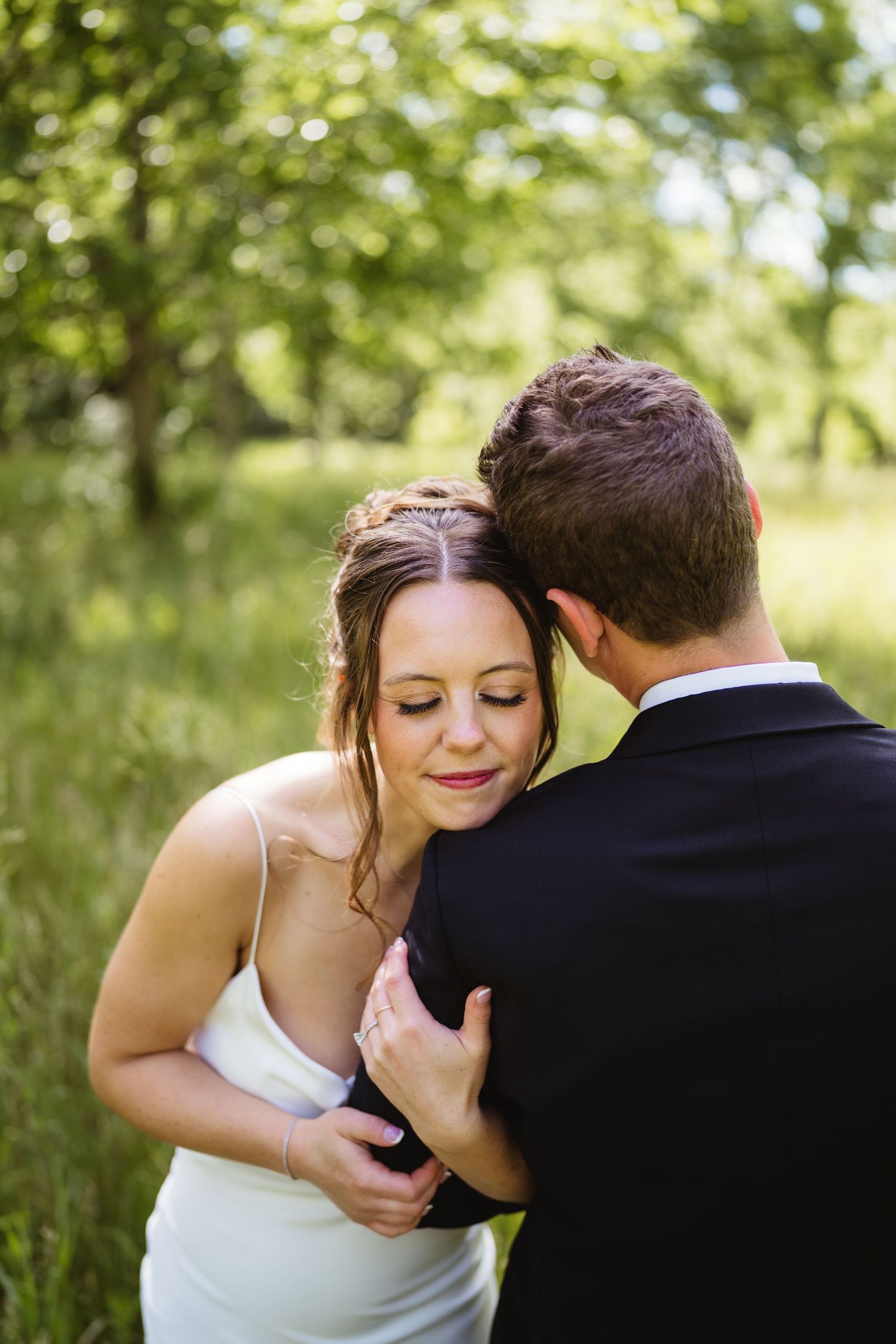 Bride and groom embrace in a lush green outdoor setting, with the bride smiling softly in a white dress.
