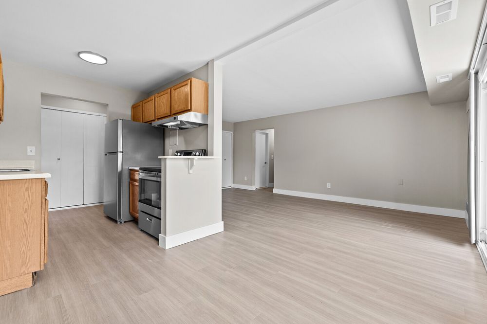 Modern apartment kitchen with stainless steel appliances, wood cabinets, and open living area beyond.