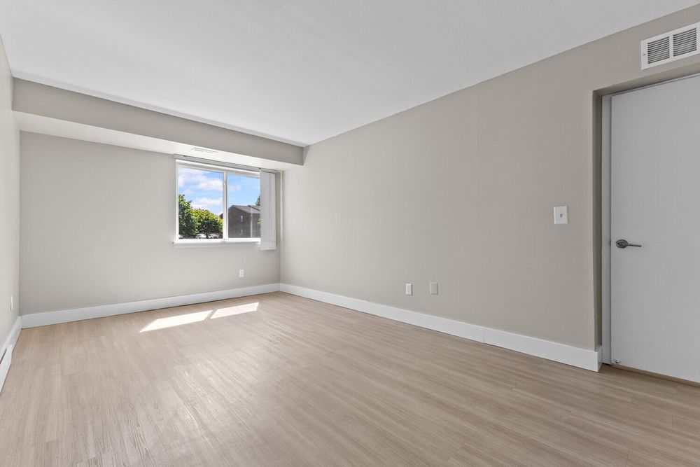Bright empty bedroom with light hardwood floors, large window, and neutral gray walls in a modern apartment.