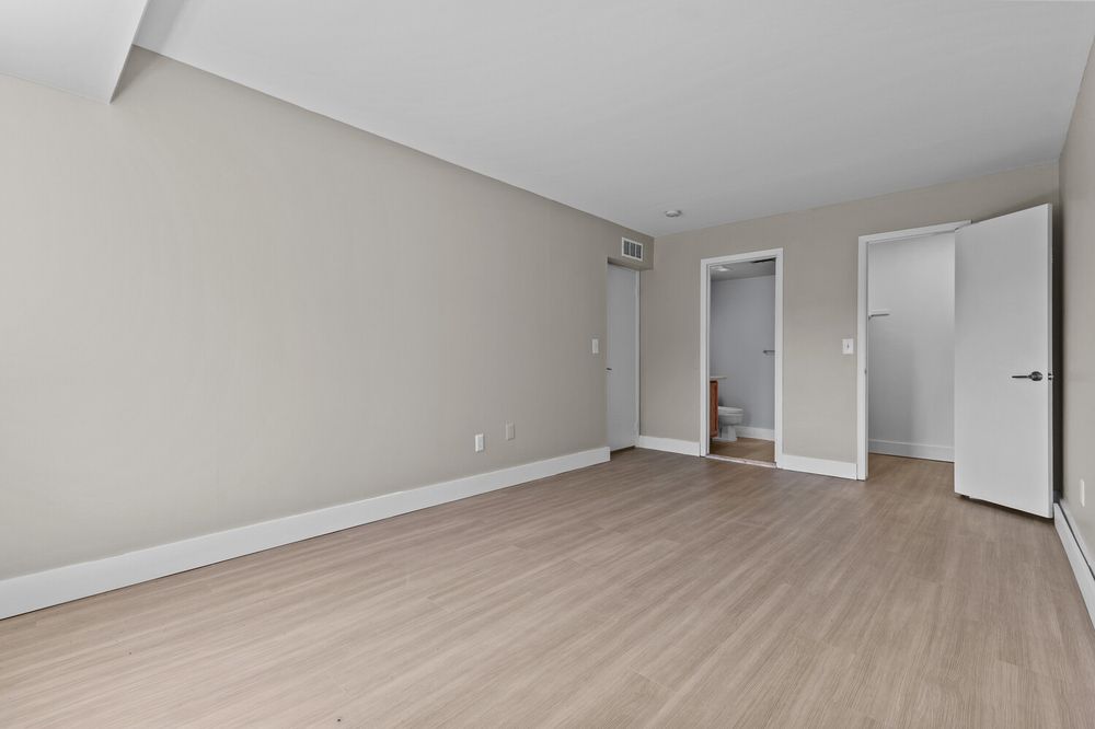 Open empty room with light hardwood floors, neutral gray walls, and doorway leading to hallway in modern apartment.