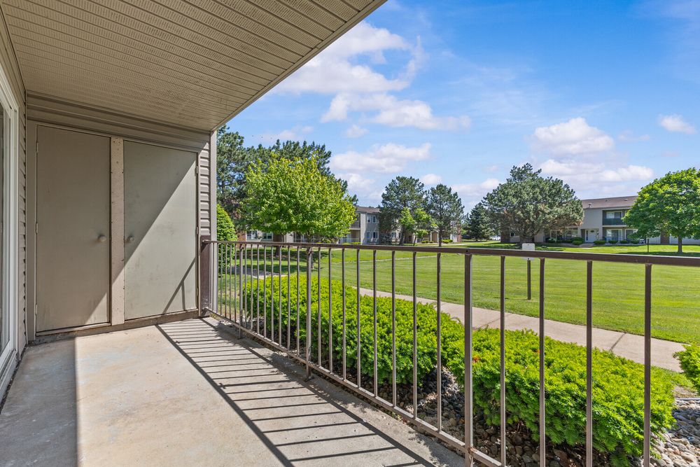Sunny apartment balcony with metal railing overlooking lush green lawn and mature trees under blue sky.