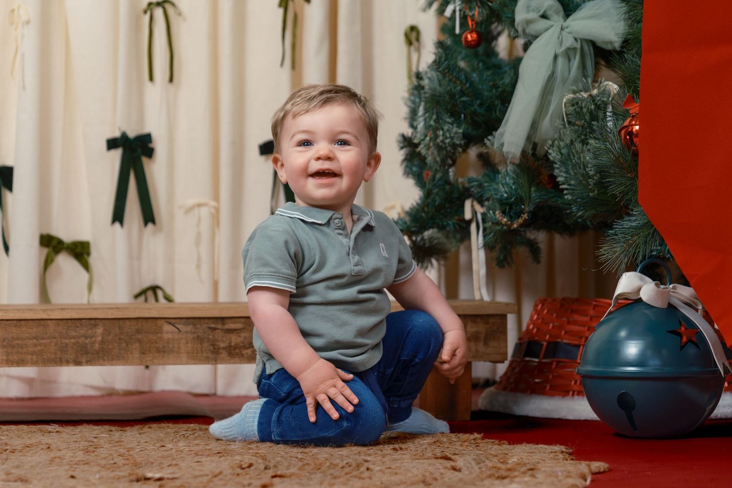 Happy toddler boy sitting on floor smiling beside a decorated Christmas tree with gifts and holiday decor.