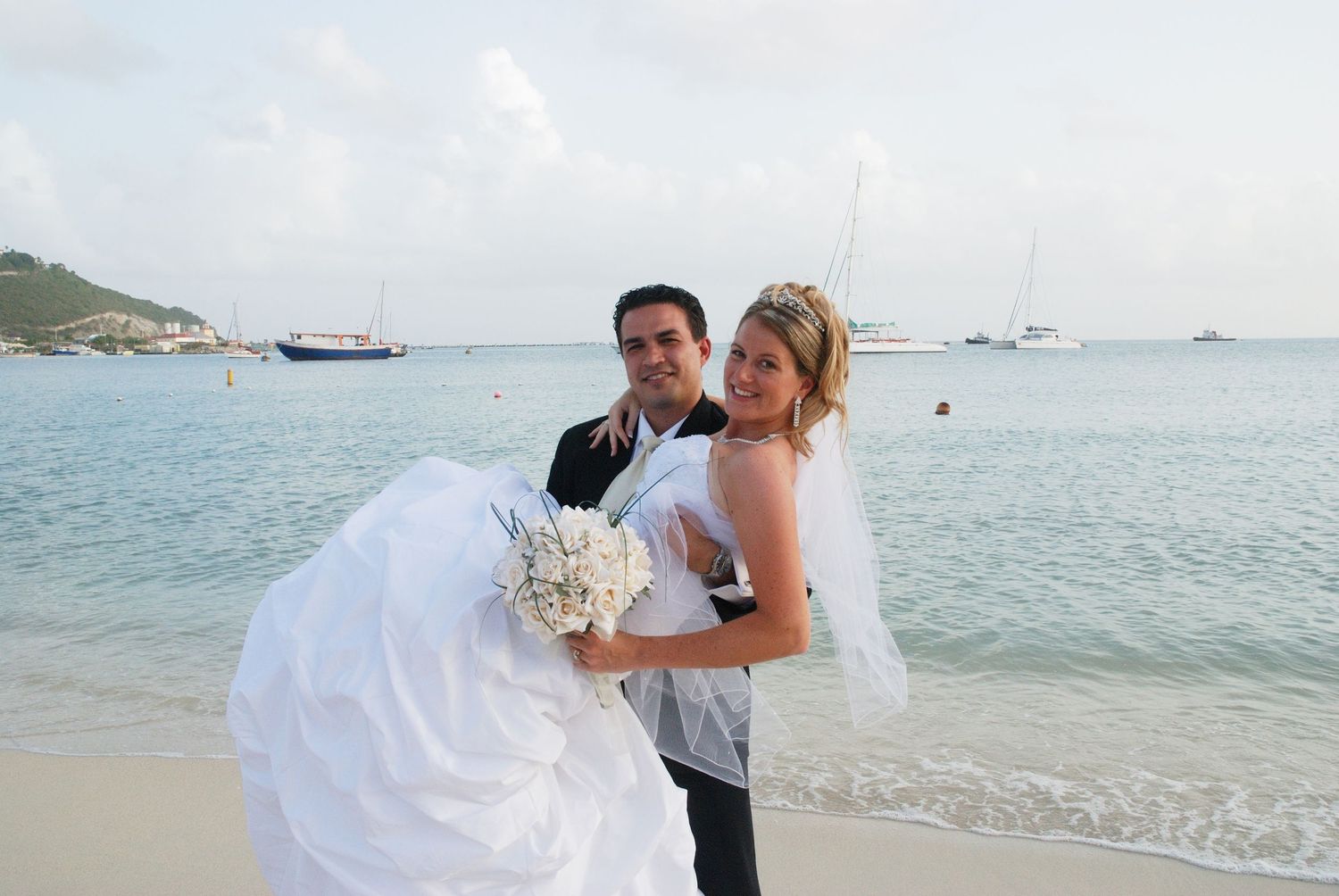 Groom carrying bride in white wedding dress on tropical beach with boats in background.
