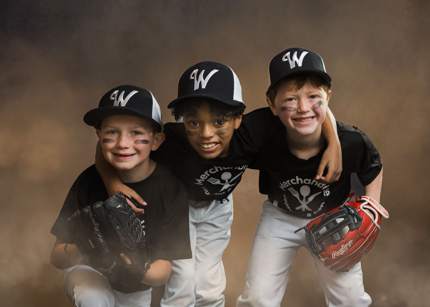 Three young boys in black Merchants baseball uniforms and caps smile together holding gloves in a team photo.