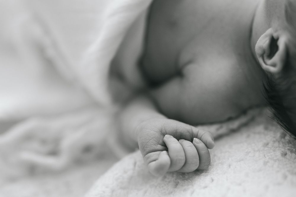 Black and white close-up of a sleeping newborn baby's tiny hand and face resting peacefully on a soft blanket.