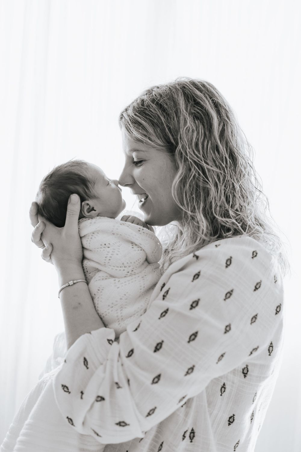 Black and white photo of a smiling mother holding her newborn baby close, touching noses in a tender moment.