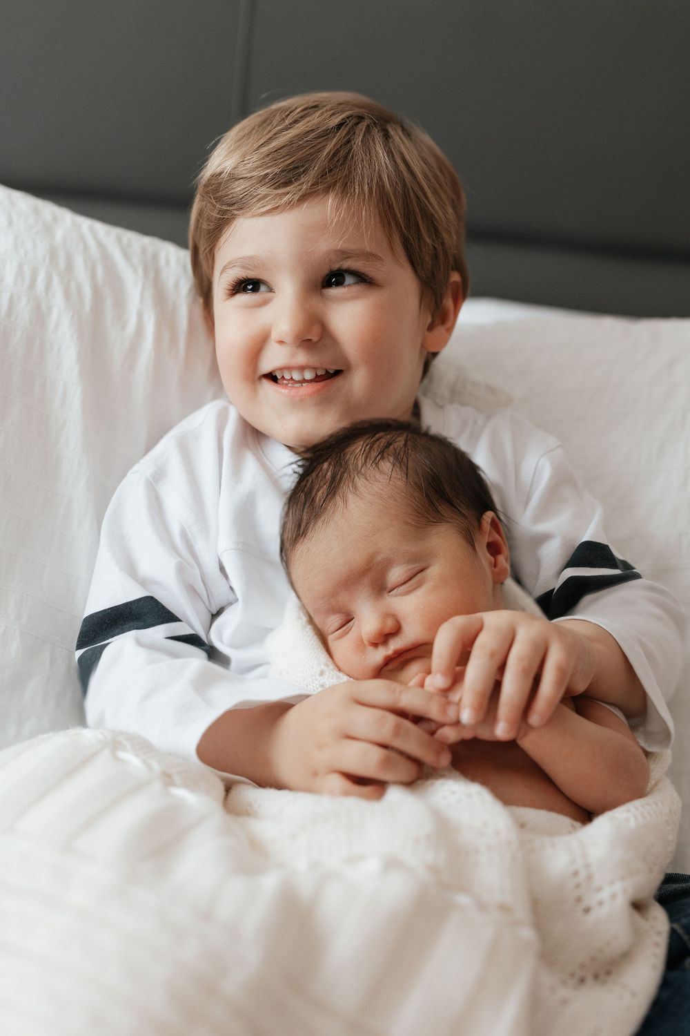 Smiling young boy holding sleeping newborn baby sibling on white bed.