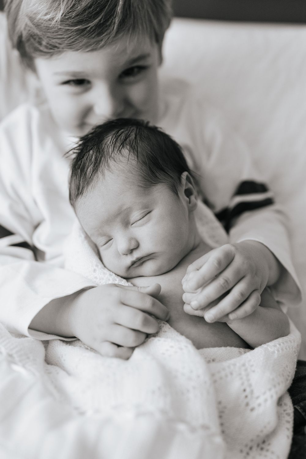 Black and white photo of a young boy gently holding his sleeping newborn sibling wrapped in a white blanket.