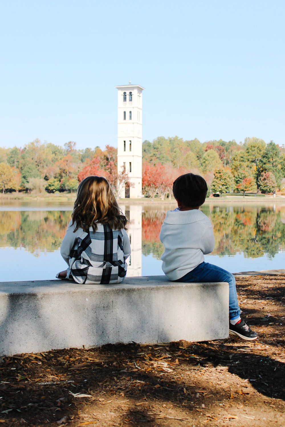 Two children sit on a stone bench overlooking a serene lake with a bell tower and colorful autumn trees reflected in the water.