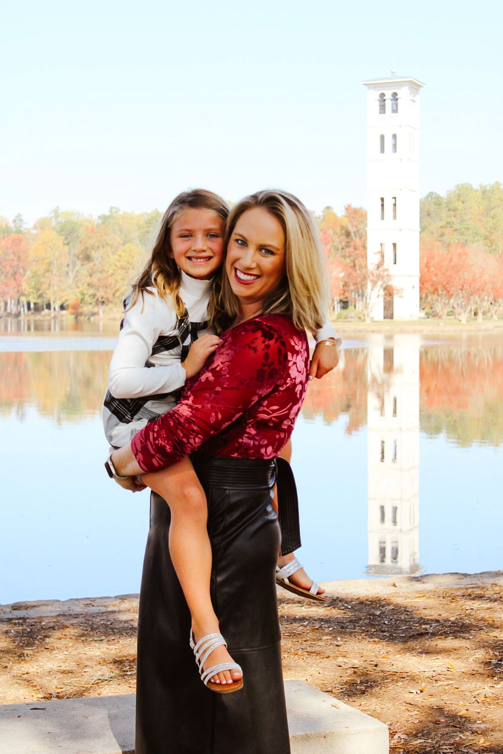 Woman in red top and black skirt holds smiling girl near a lake with a white bell tower in autumn.