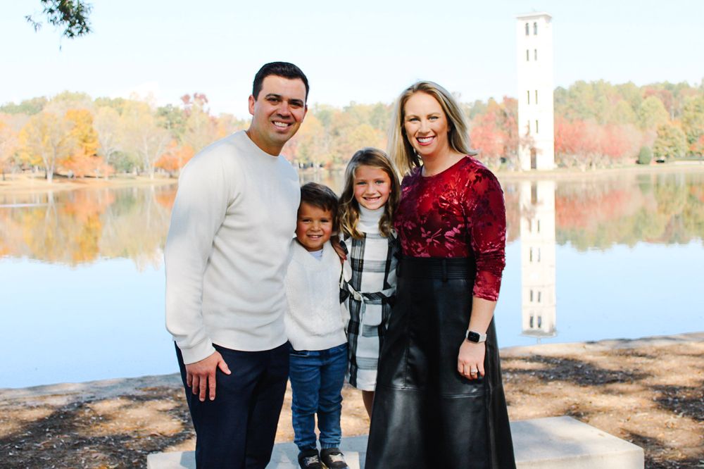 Family of four smiling outdoors by a lake with autumn foliage and a tower in the background.
