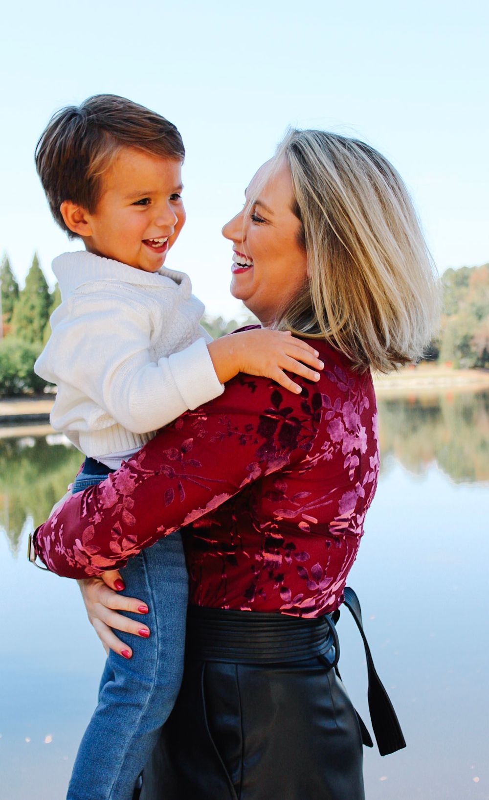 A joyful mother in a red floral top holds her laughing young son near a serene lakeside setting.