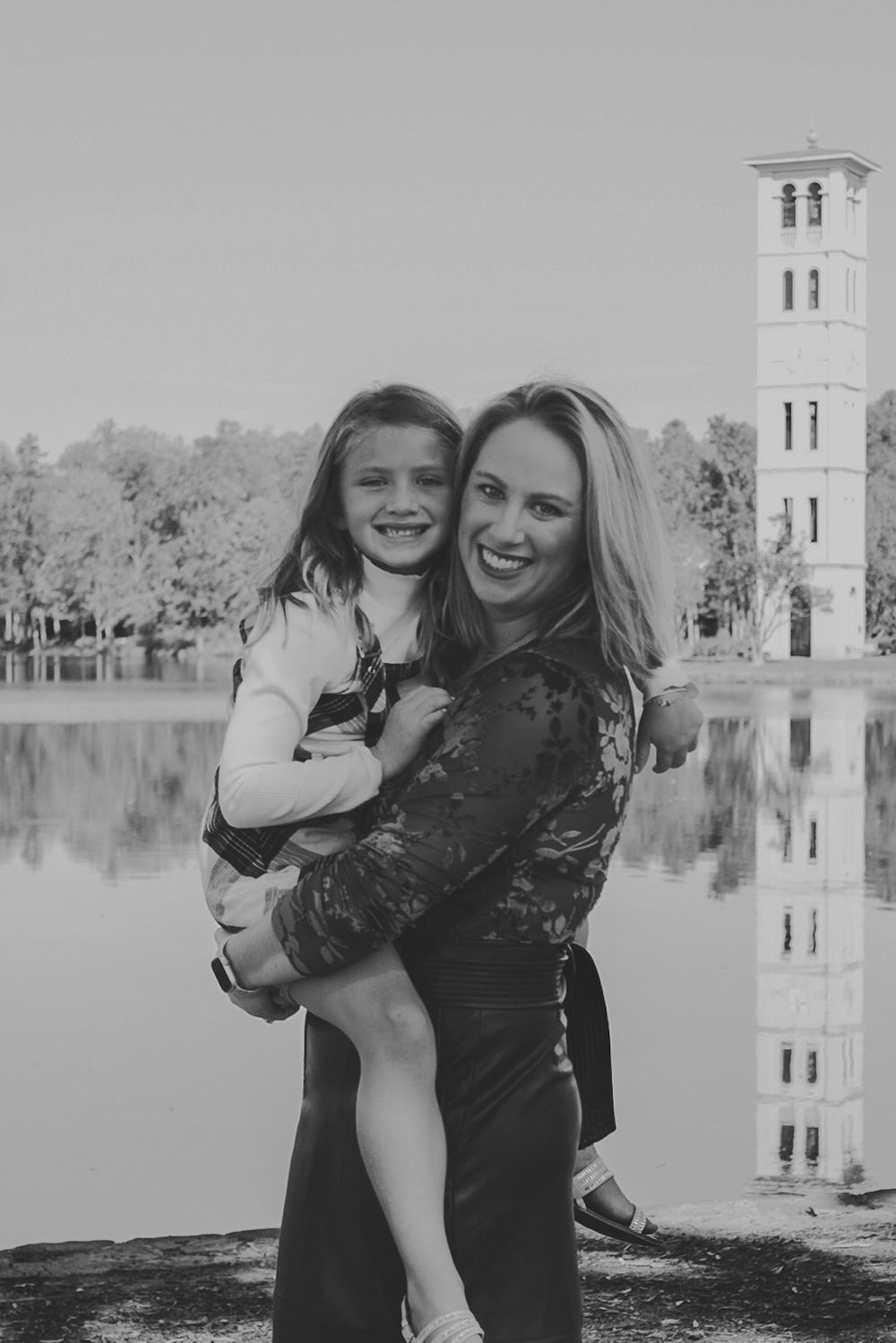 Black and white photo of a smiling mother holding her young daughter near a lake with a tower reflected in the water.