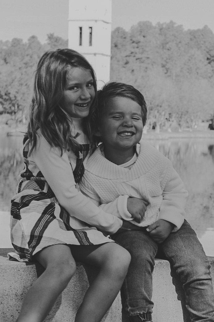 Two smiling children sit together outdoors near a lake and tower in a black and white portrait photo.