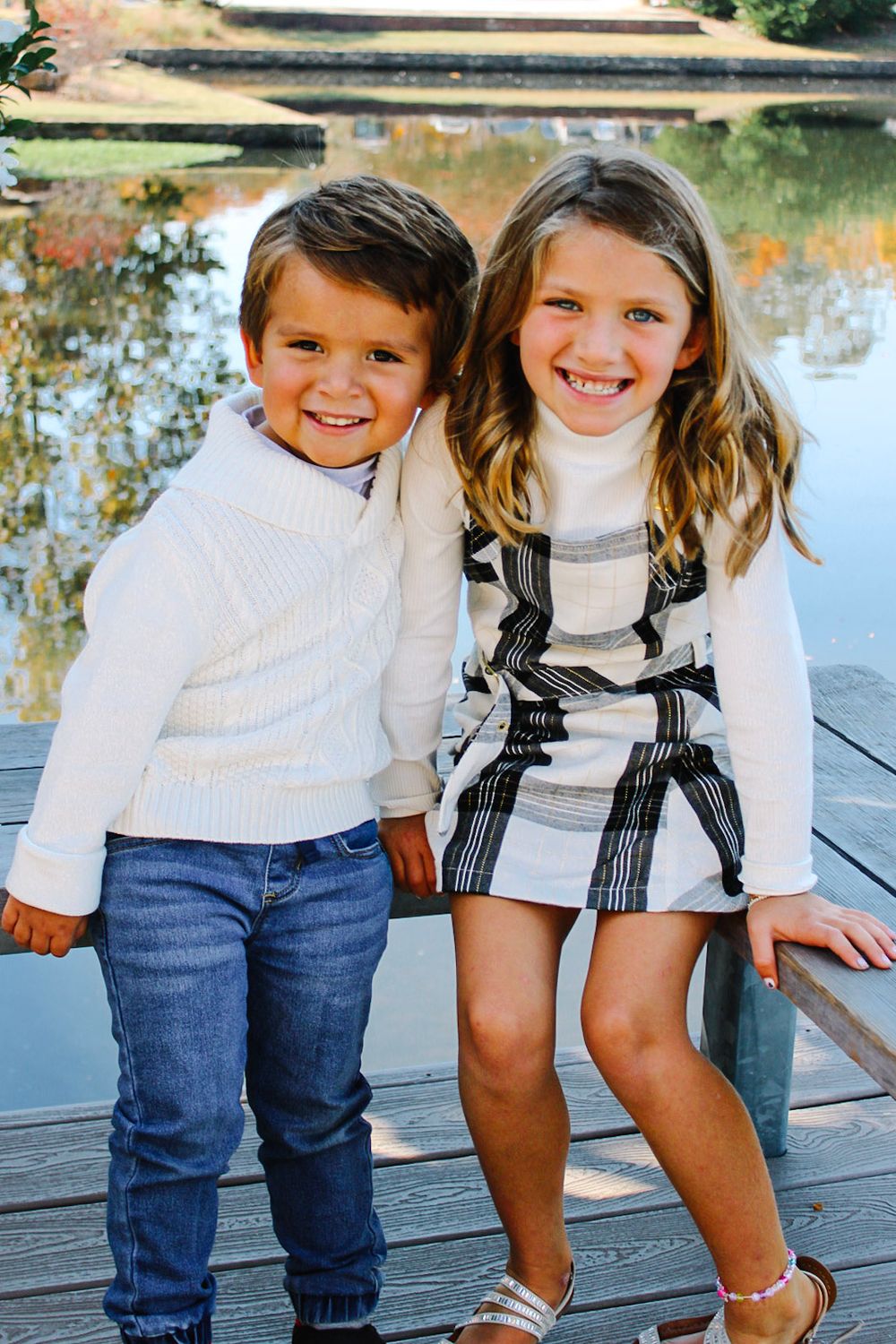 Two smiling children pose on a wooden dock by a pond, boy in white sweater and jeans, girl in plaid dress.