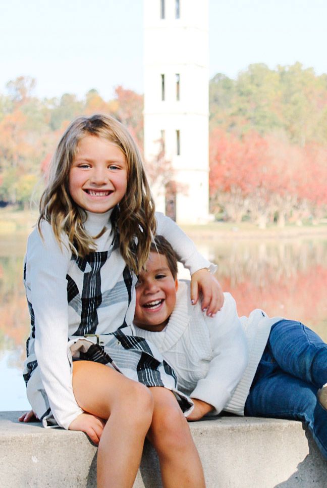Two smiling children sit together outdoors near a lake with fall foliage and a tower in the background.