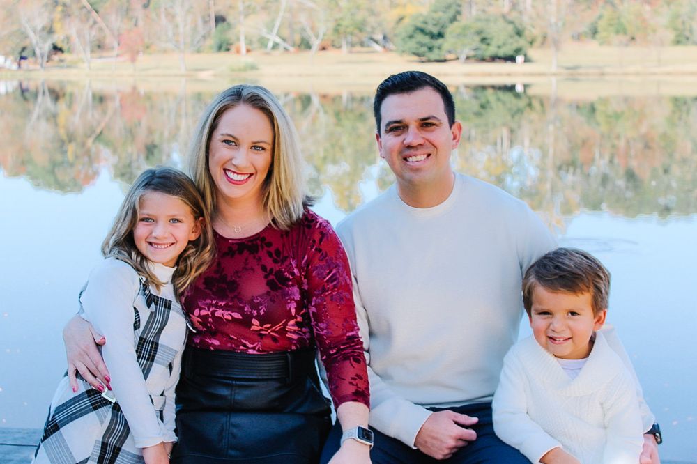 Happy family of four smiling together outdoors by a serene lake on a sunny day.