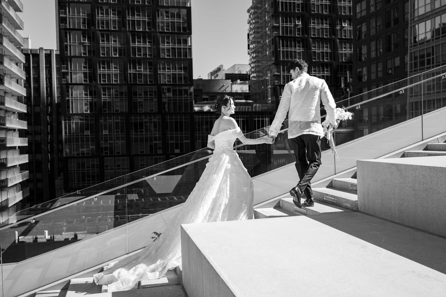Black and white photo of a bride and groom holding hands walking up outdoor stairs with city buildings in background.