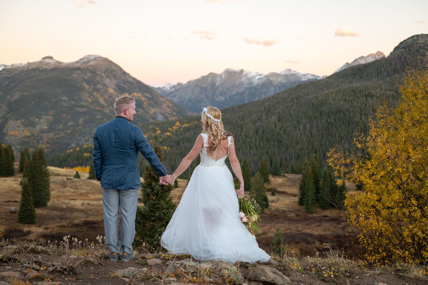 A bride and groom stand in the San Juan mountains. They are facing away from the camera but looking at each other.