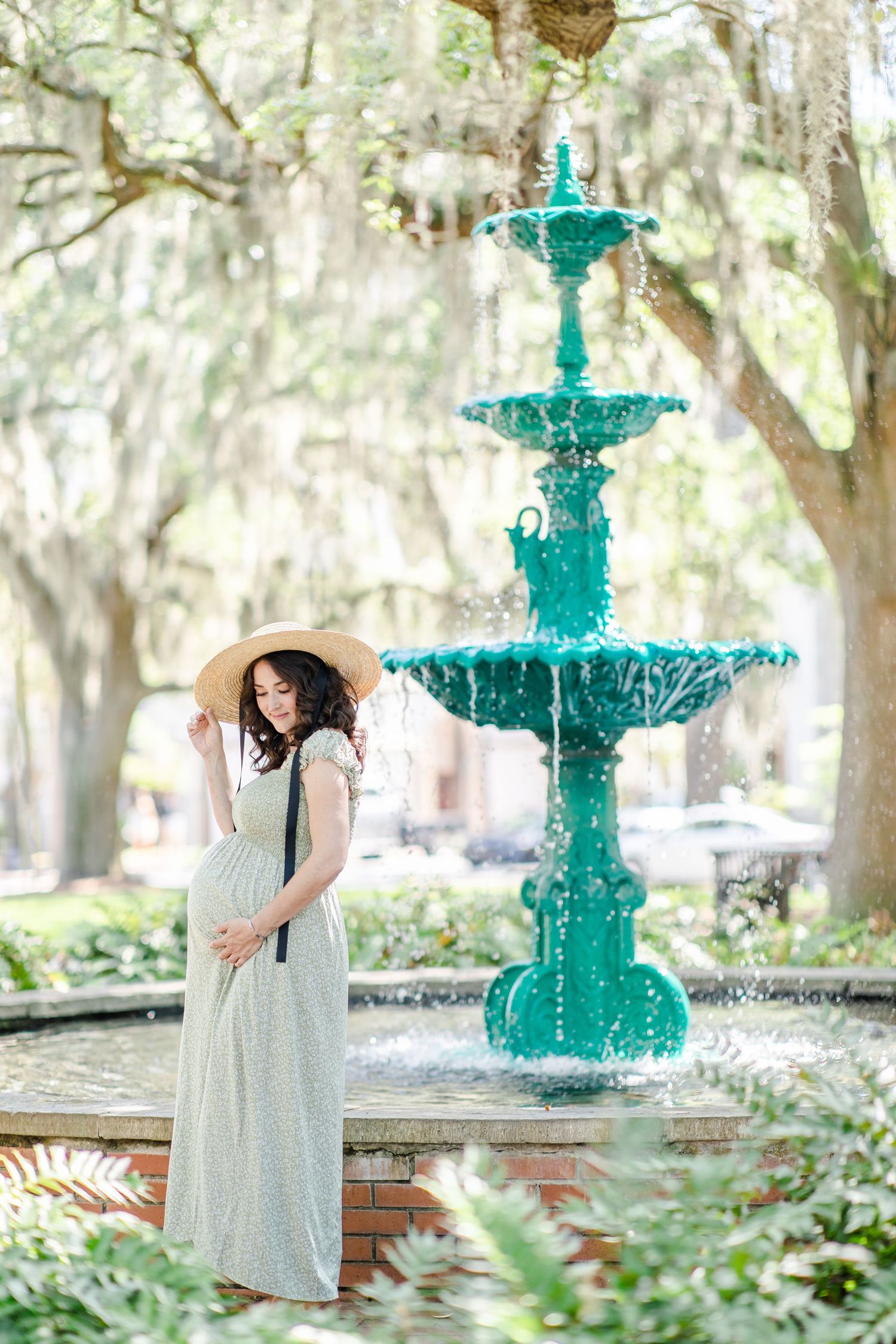 Pregnant woman in mint dress and sun hat poses by a turquoise tiered fountain in a sunny park.