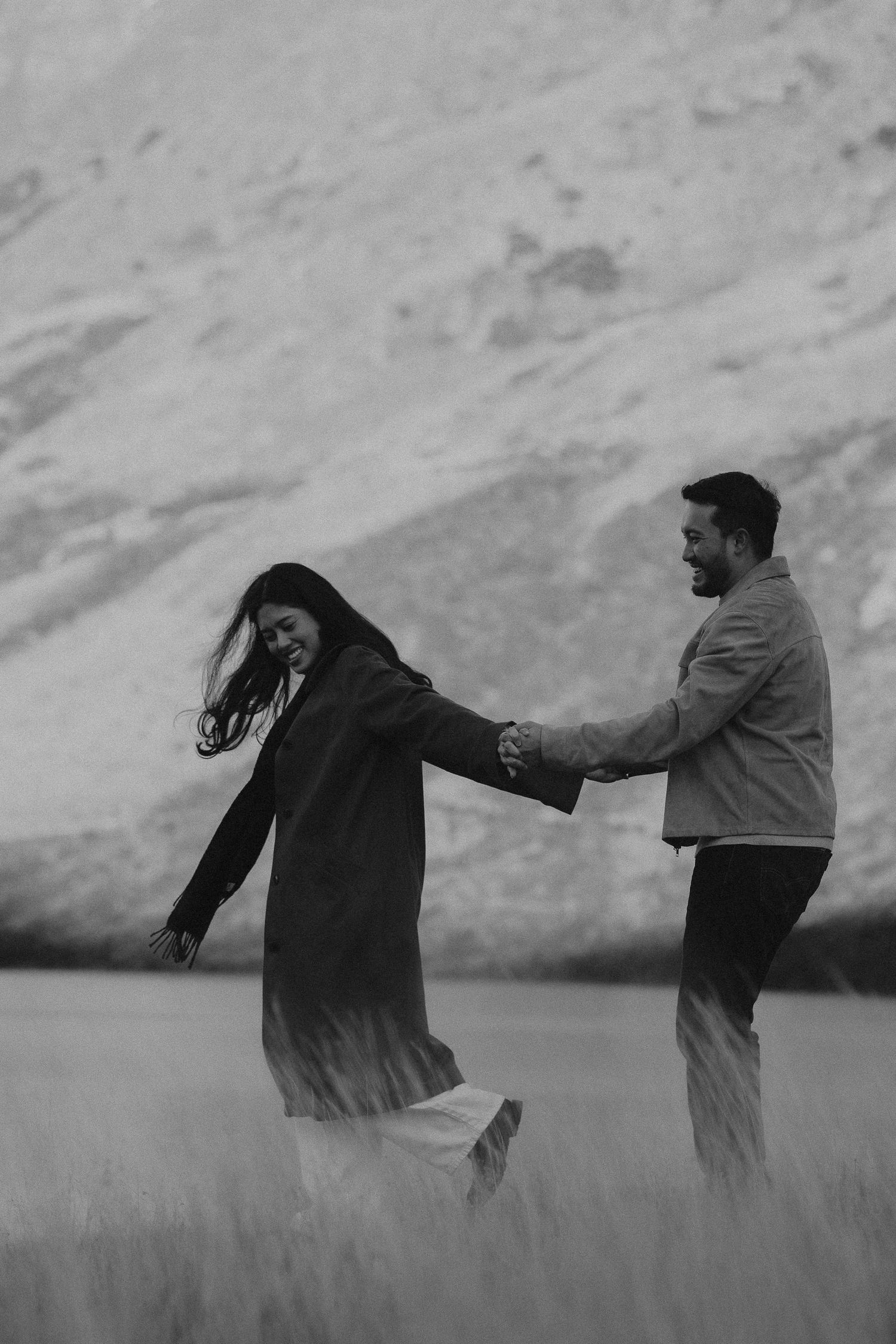 Black and white photo of a joyful couple holding hands and dancing in a field with a mountain backdrop.