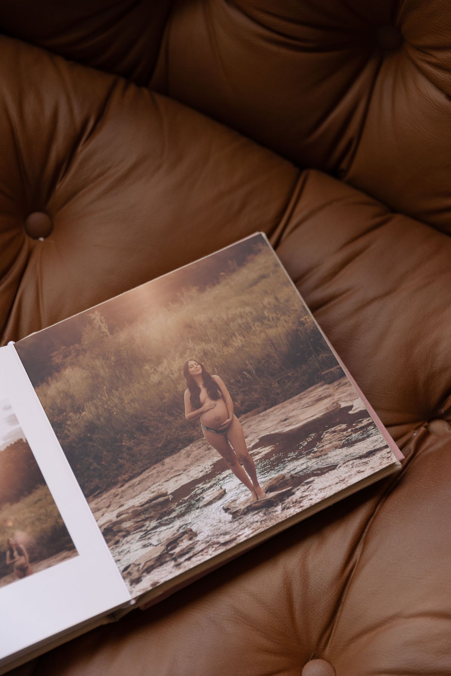 Open photo book on brown tufted leather sofa displaying a woman standing on rocks in a sunlit forest setting.