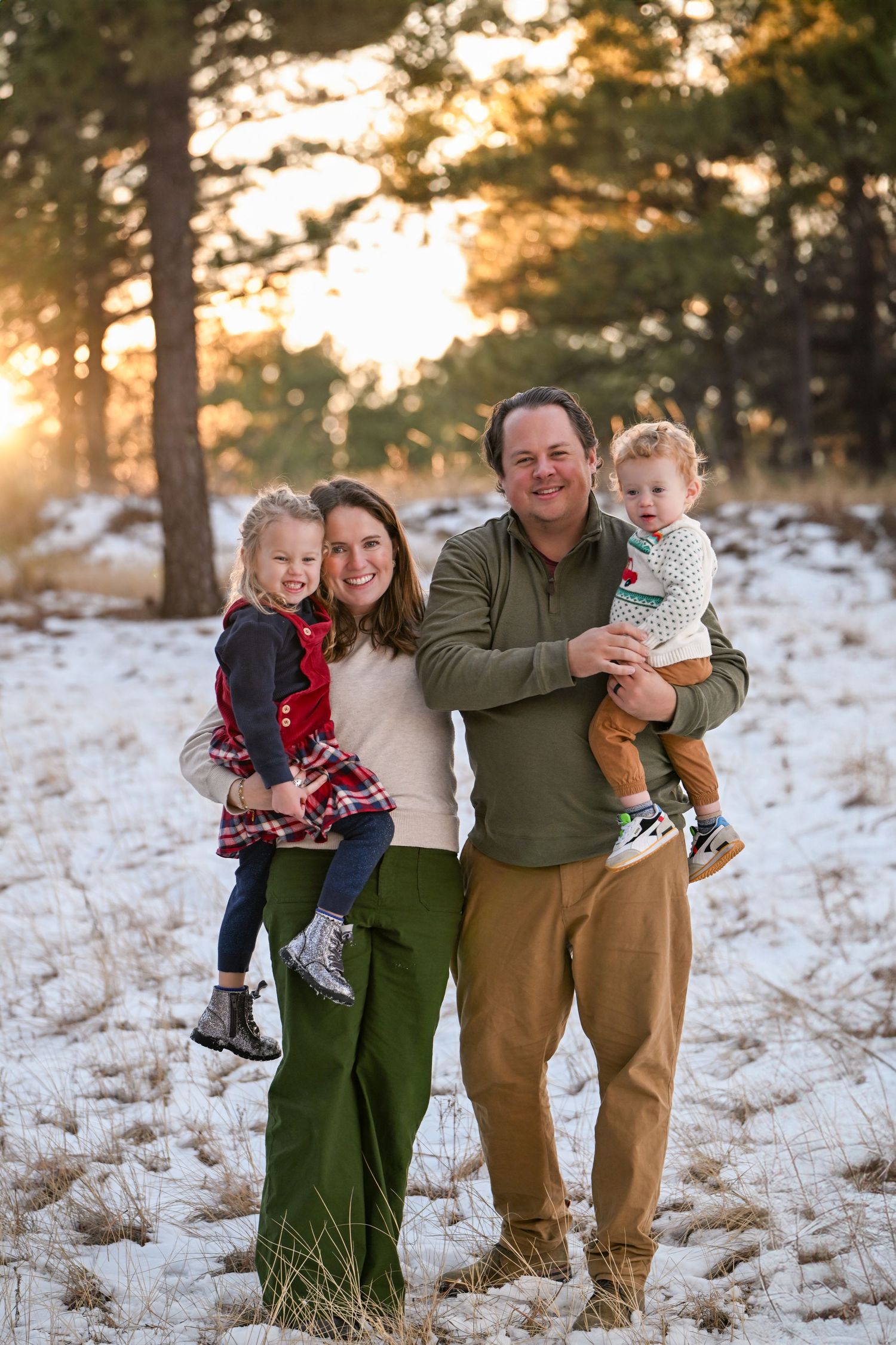 Family of four smiling in snowy pine forest at golden hour, parents holding two young children in holiday outfits.