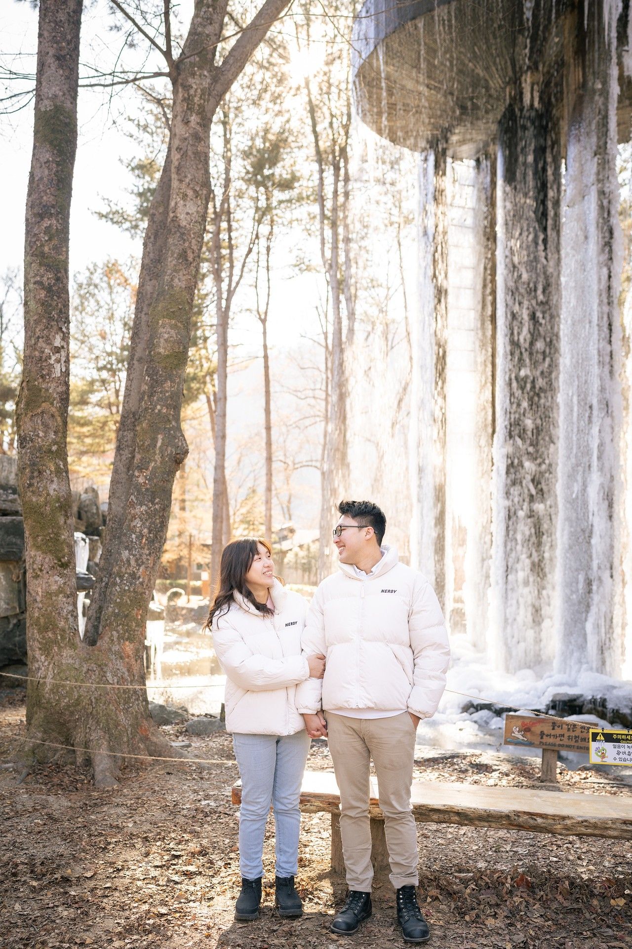 Couple in white puffer jackets holding hands before a frozen waterfall in a winter forest.