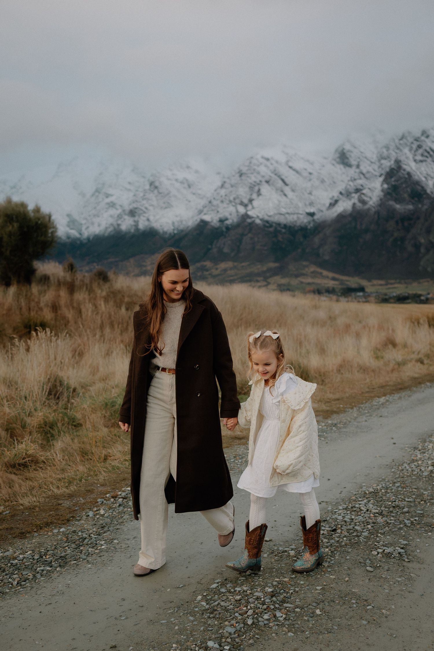 A woman and young girl walk hand-in-hand on a gravel path with snow-capped mountains in the background.