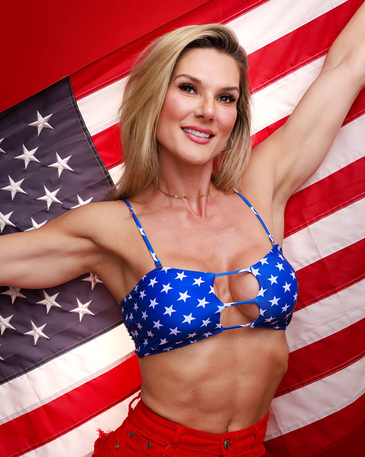 Blonde woman in white cowboy hat draped in American flag smiling against red background.
