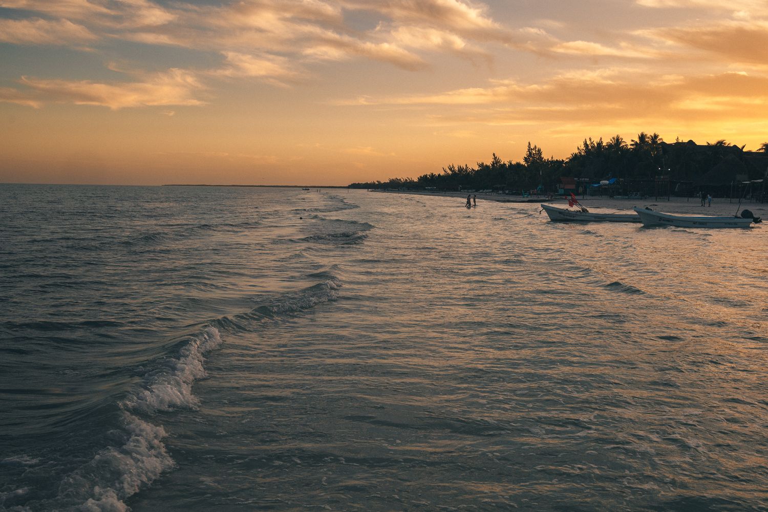 Golden sunset over tropical beach with gentle waves and boats along the shoreline.