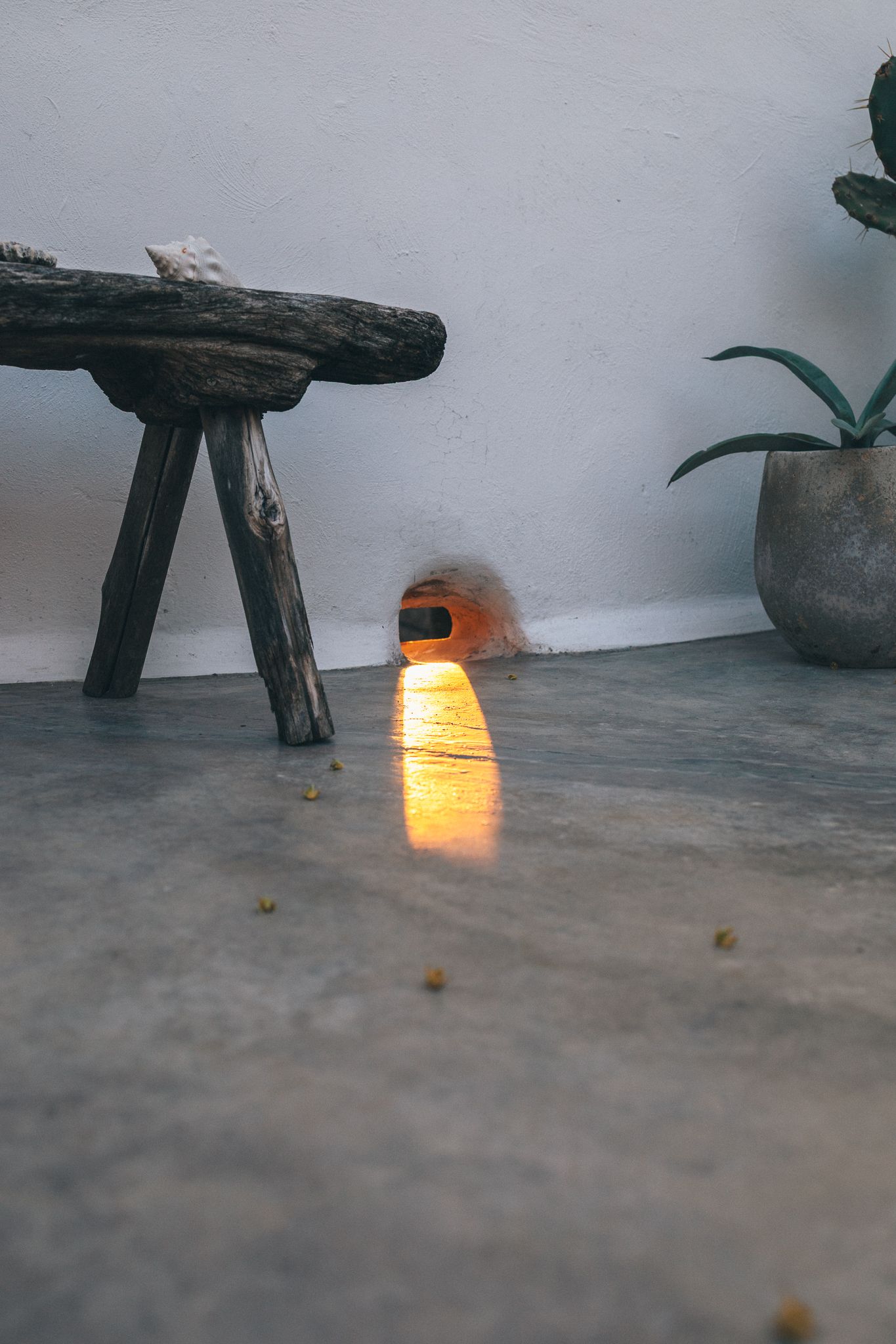 Small orange glowing floor lamp beside rustic wooden stool and potted plant on concrete floor.
