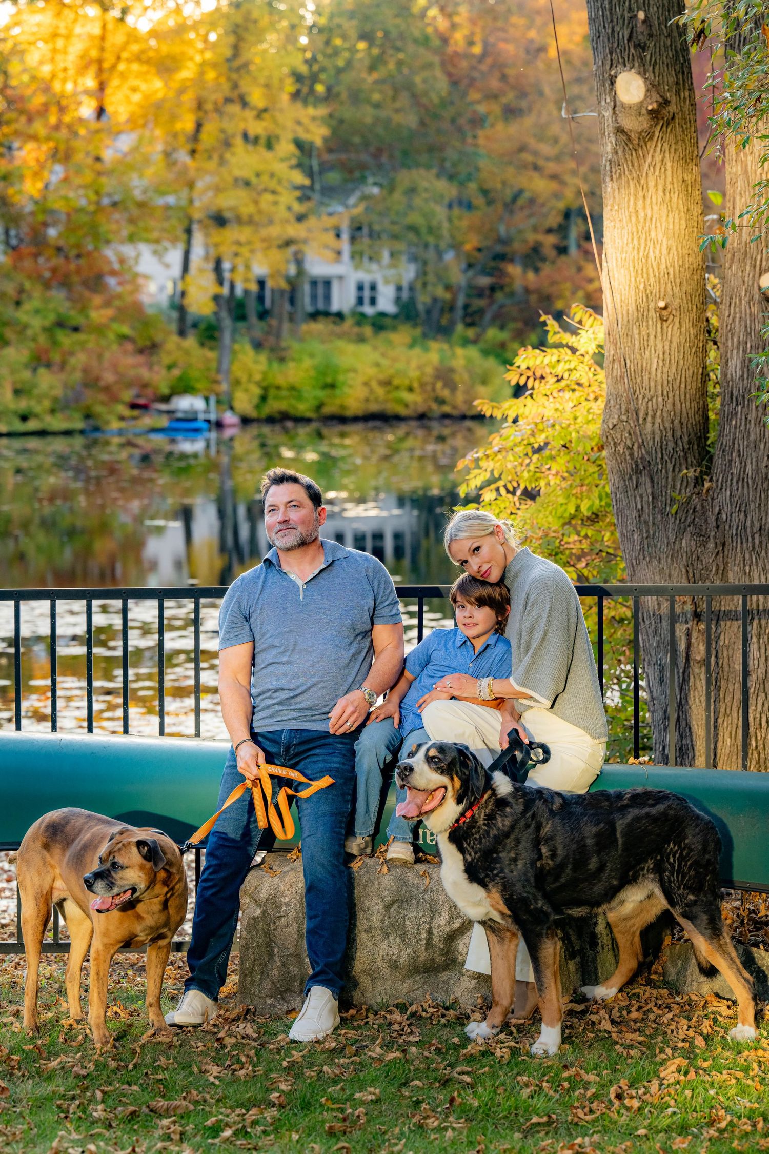 Family with two dogs posing by a lakeside park bench surrounded by colorful autumn foliage.