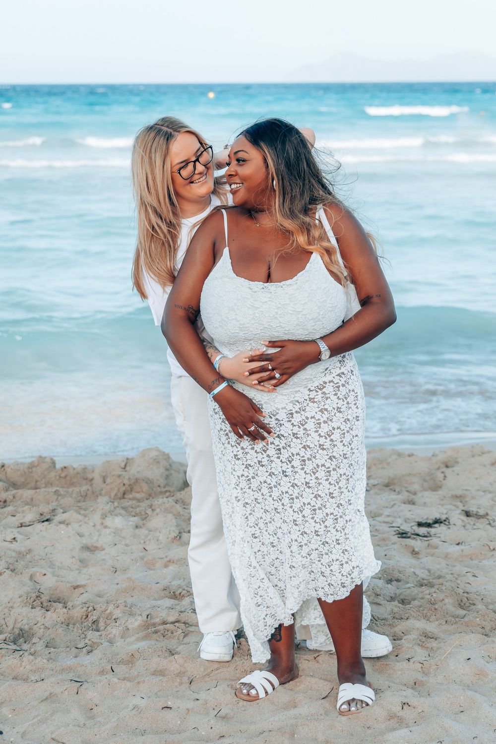 An elegant gay couple embracing from behind and smiling on the shore during a surprise proposal photoshoot in Mallorca.