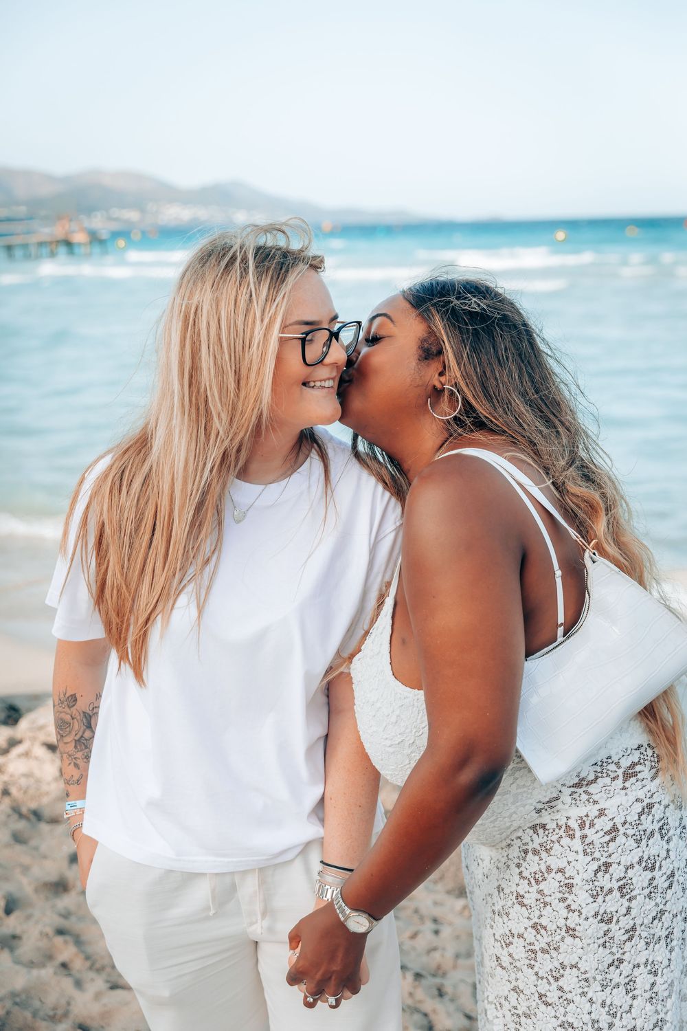 Two women sharing an affectionate embrace and a kiss on the cheek during a romantic LGBTQ+ engagement session on a Mallorca beach.