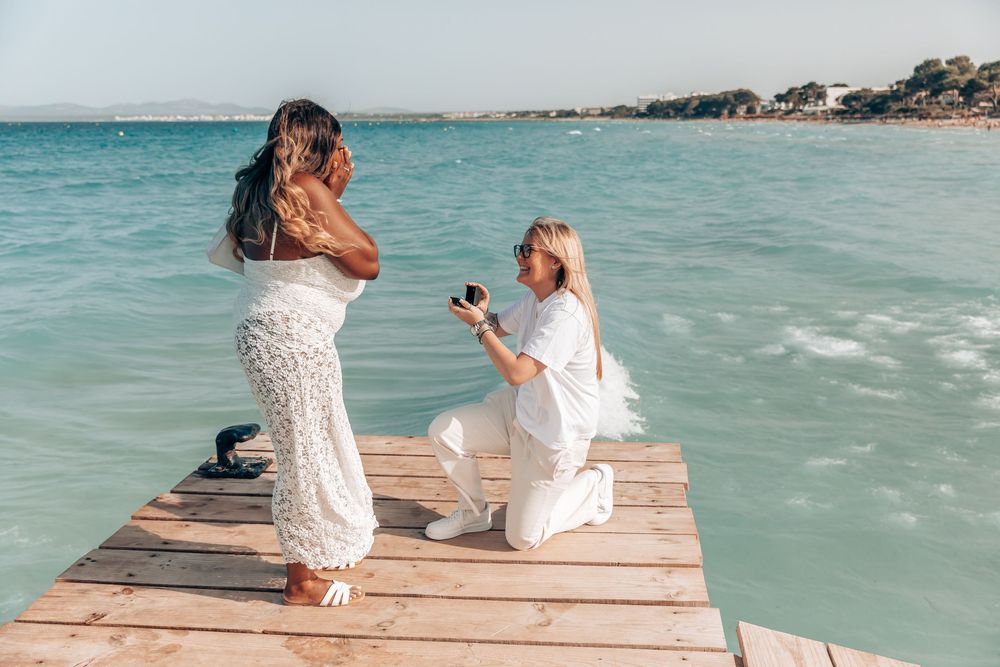 A woman down on one knee proposing to her girlfriend on a rustic wooden pier during a surprise Mallorca beach proposal.