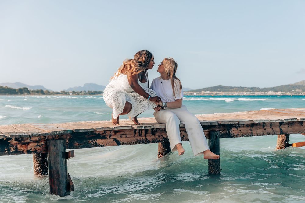 Wide shot of a lesbian couple sharing an intimate moment while sitting together on a wooden pier above turquoise water in Mallorca.
