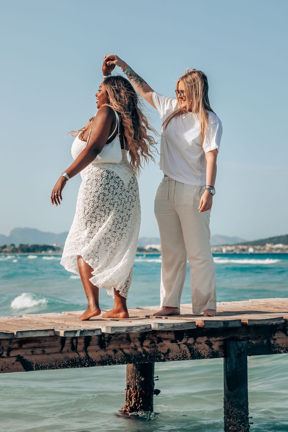 Two women in matching white outfits laughing and dancing together on a rustic wooden pier during their Mallorca engagement session.
