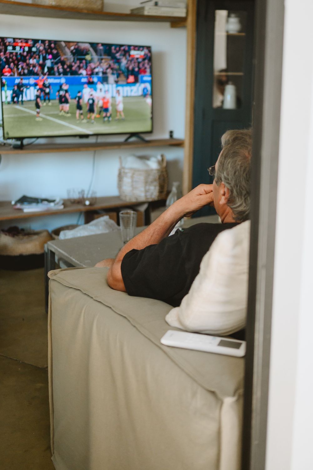 Elderly man watching a soccer match on a large flat screen TV from a cozy sofa.