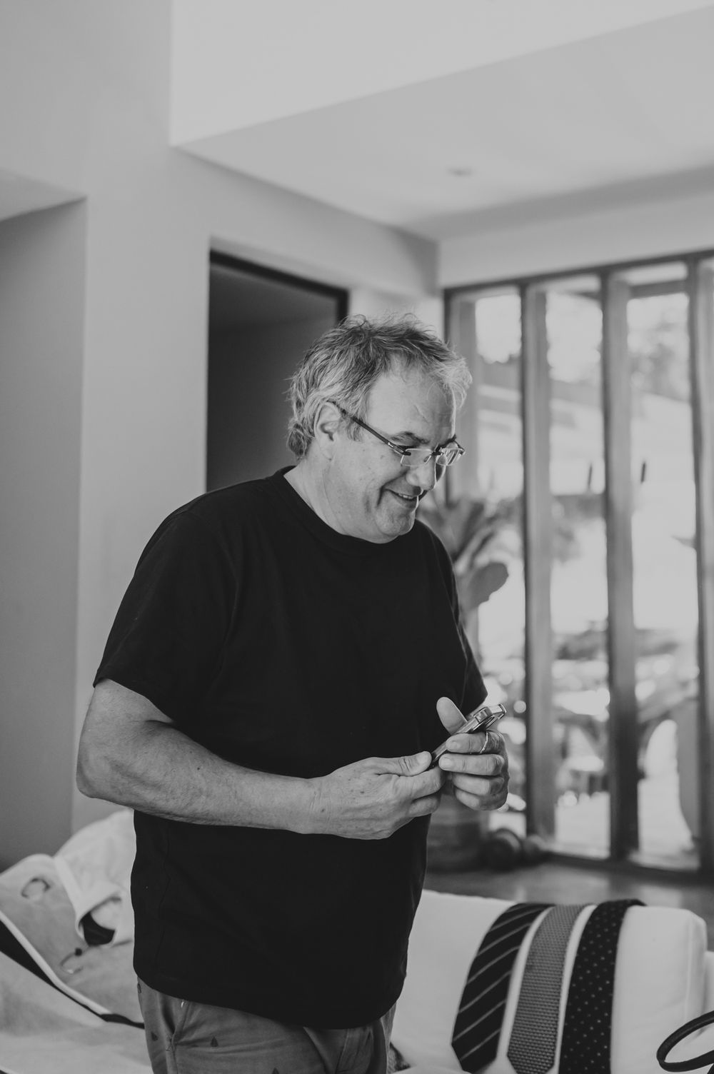 Black and white photo of a smiling middle-aged man in a black t-shirt holding cufflinks.