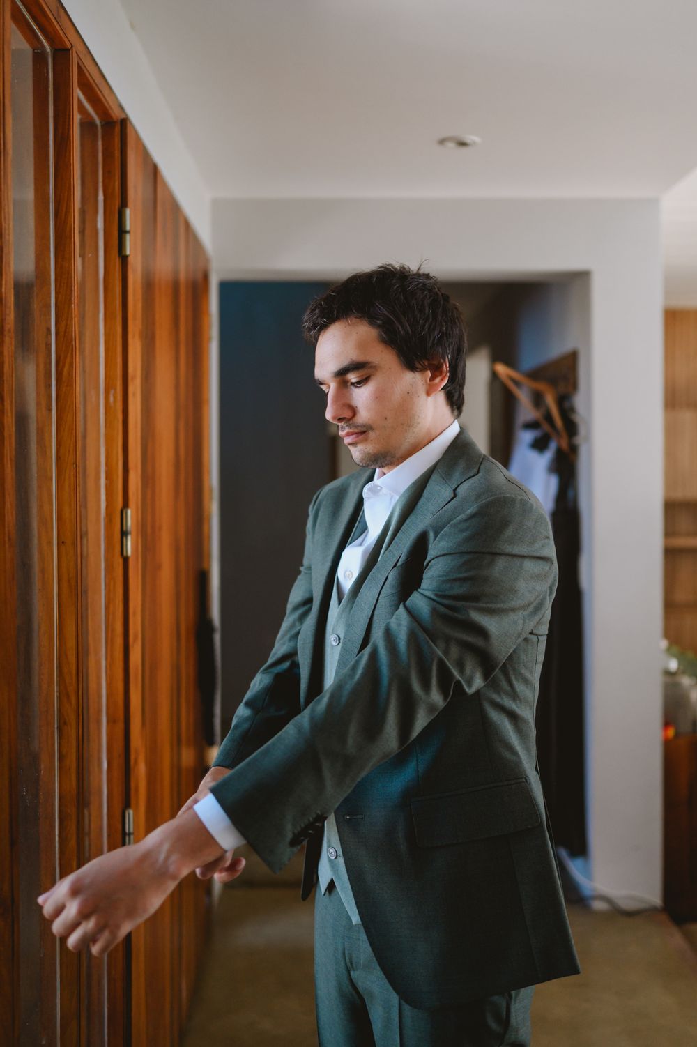 Young man in dark green three-piece suit adjusting his cufflinks indoors.