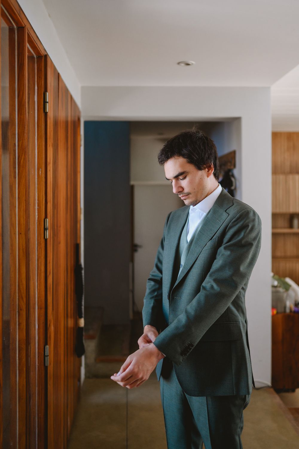Groom in elegant dark green three-piece suit adjusting his sleeve before wedding.
