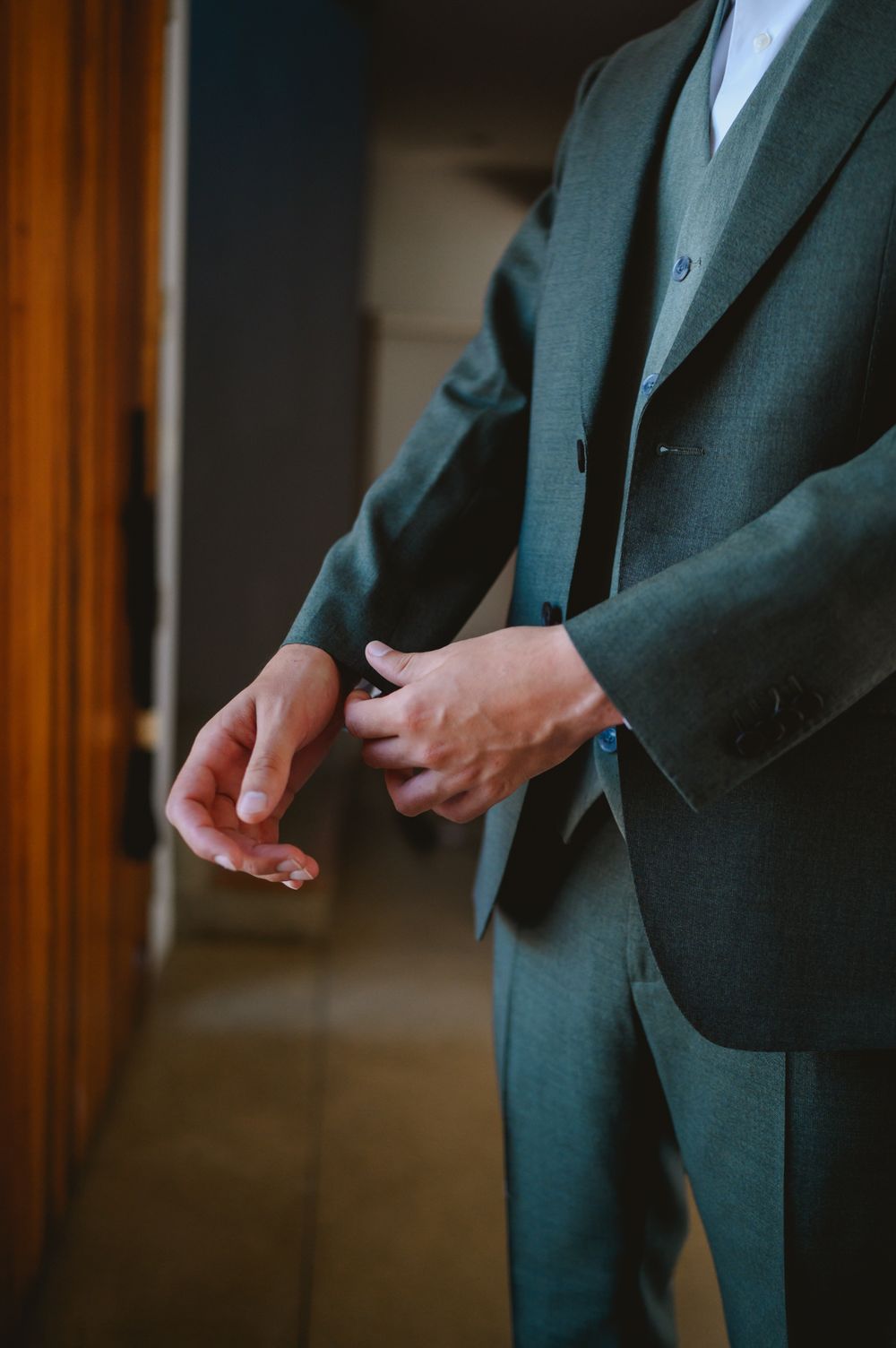 Close-up of groom fastening buttons on dark green suit jacket before ceremony.