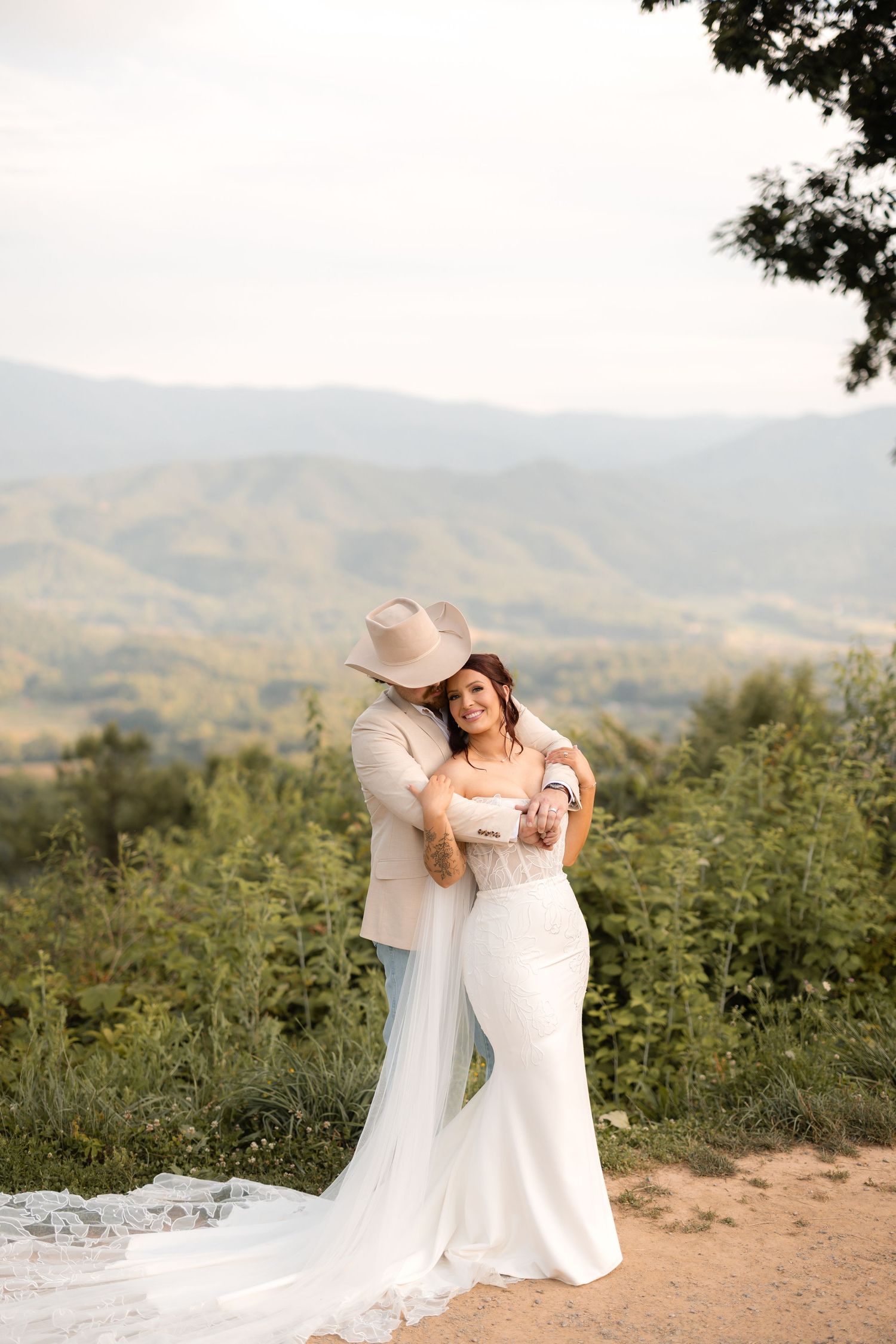 Bride and groom embracing outdoors in mountain landscape, wearing white gown and tan cowboy hat.